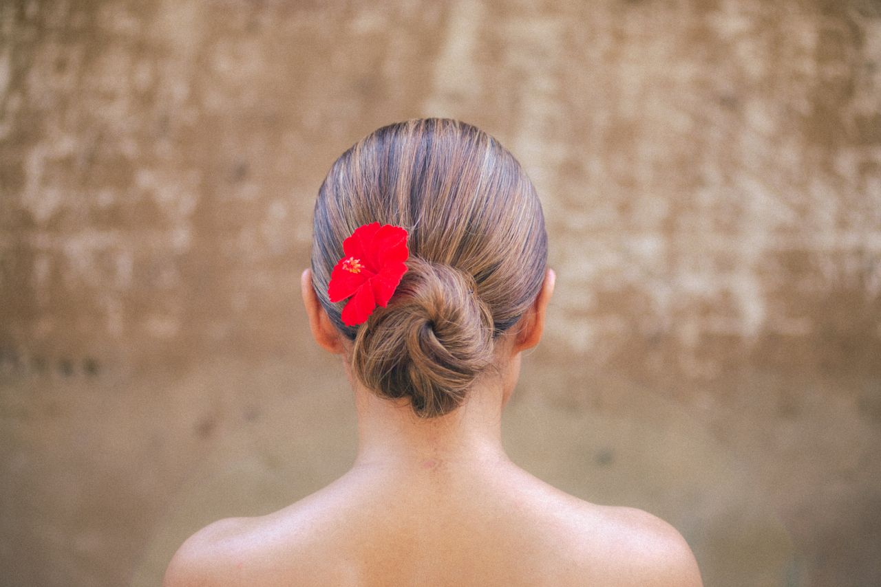 Back view of a woman with a red flower in her hair bun against a rustic backdrop, Bali.